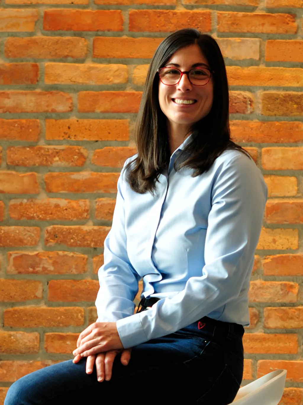 Ágnes Farkas is wearing glasses and a blue blouse and is smiling at the camera in front of a wall.