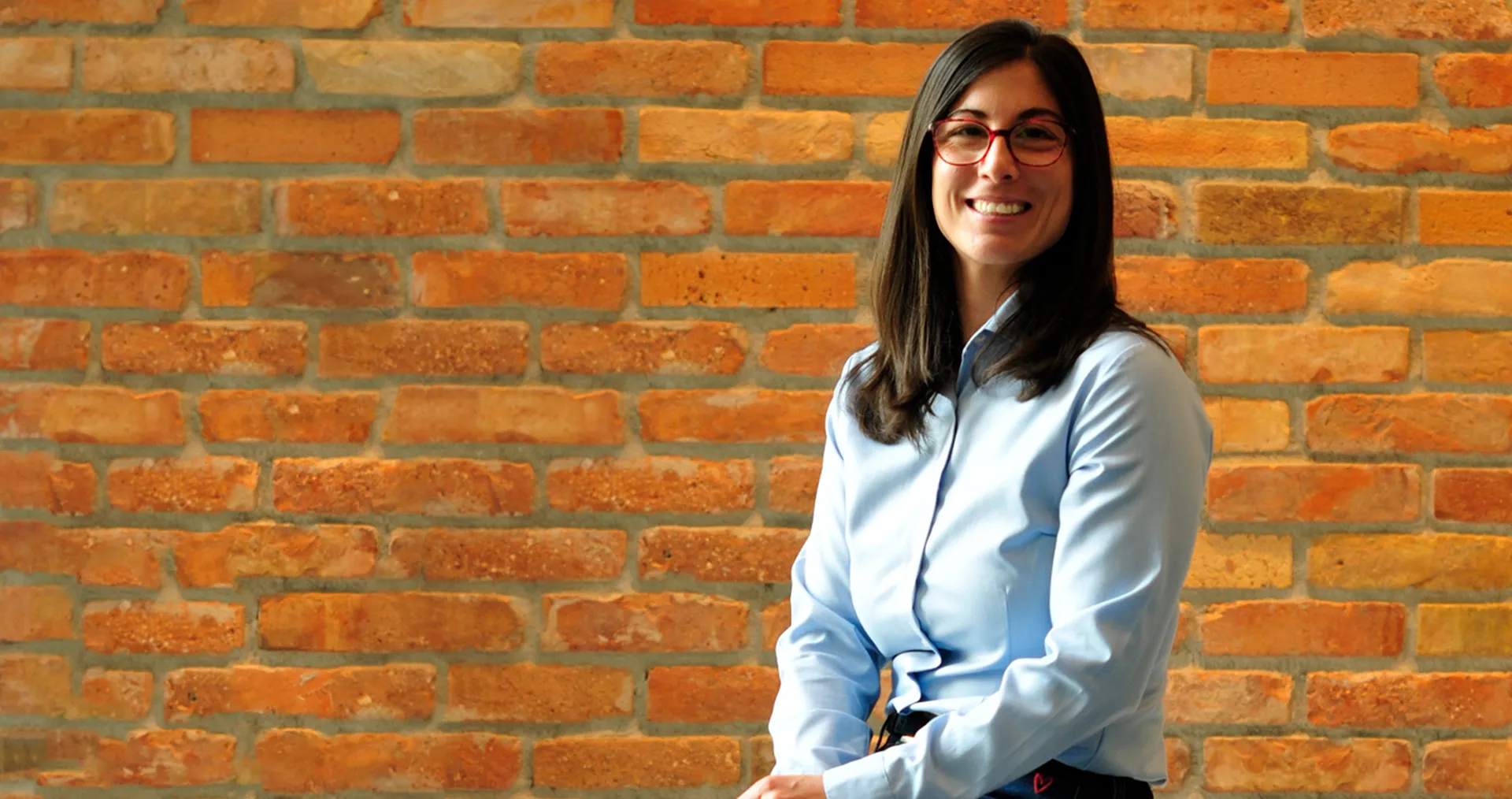 Ágnes Farkas is wearing glasses and a blue blouse and is smiling at the camera in front of a wall. (Photo)