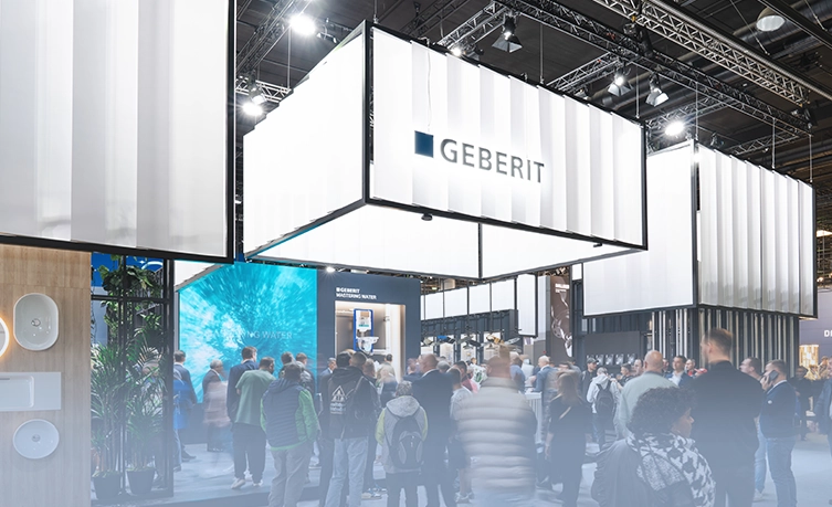 The image shows an exhibition with a modern and geometric architectural design. There are people gathered around booths and information stands. There's a clear sign that says "Geberit", the company being represented at one of the booths. (Photo)