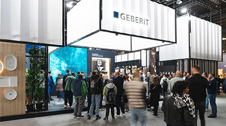 The image shows an exhibition with a modern and geometric architectural design. There are people gathered around booths and information stands. There's a clear sign that says "Geberit", the company being represented at one of the booths. (Photo)